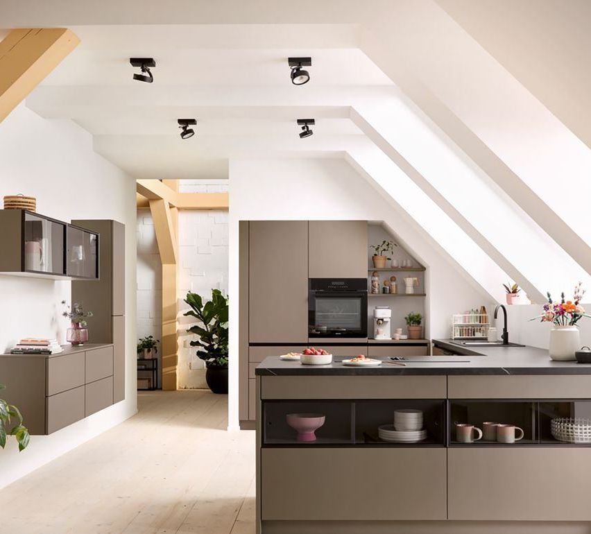A modern kitchen with a large island and cabinets, a microwave, and plants on the counters. The ceiling has sloped beams and recessed lighting.