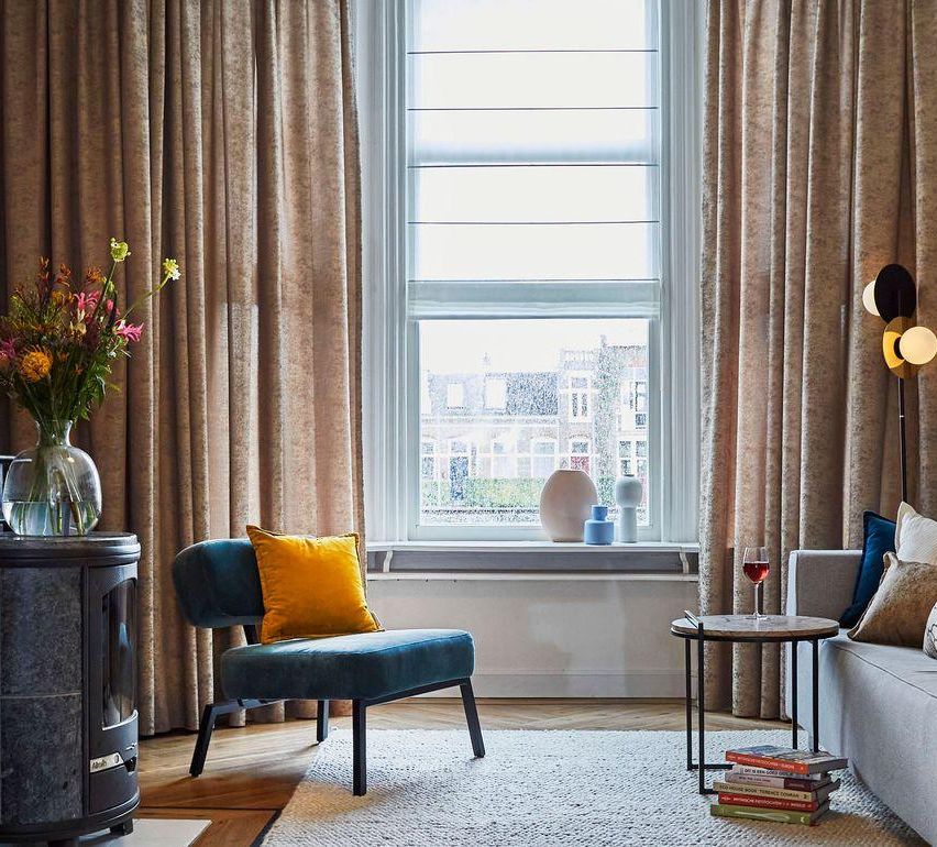 A cozy living room with a blue chair and a yellow pillow, a window with curtains, a small table with a wine glass, and a couch with pillows.