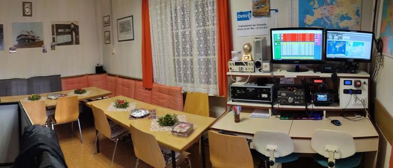 Ein Innenansicht eines Raumes mit einem langen Tisch und Stühlen, einer Couch und mehreren Bildern an der Wand. Ein Monitor befindet sich auf der rechten Seite, mit einem Regal darunter. Eine Leiter steht an der Wand auf der linken Seite.