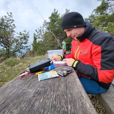 Ein Mann sitzt auf einer Bank im Freien, trägt eine Winterjacke und schreibt in ein Notizbuch. Verschiedene Gegenstände wie ein Radio, Snacks und eine Flasche stehen auf dem Tisch vor ihm.