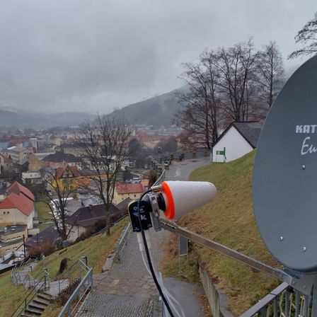Ein erhöhter Blick auf eine Stadt mit einer Satellitenschüssel und Kabel auf der Seite eines Hügels. Im Hintergrund sind Gebäude, Bäume und Berge zu sehen.