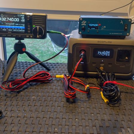 Ein Funkgerät mit einem digitalen Display, das 432.740.00 anzeigt, steht auf einem Stativ. Ein Netzteil mit einem Display, das 14.80 anzeigt, ist mit Kabeln verbunden.