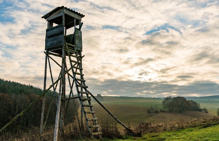 Ein alter hölzerner Aussichtsturm auf einem grünen Hang mit einem bewölkten Himmel im Hintergrund.