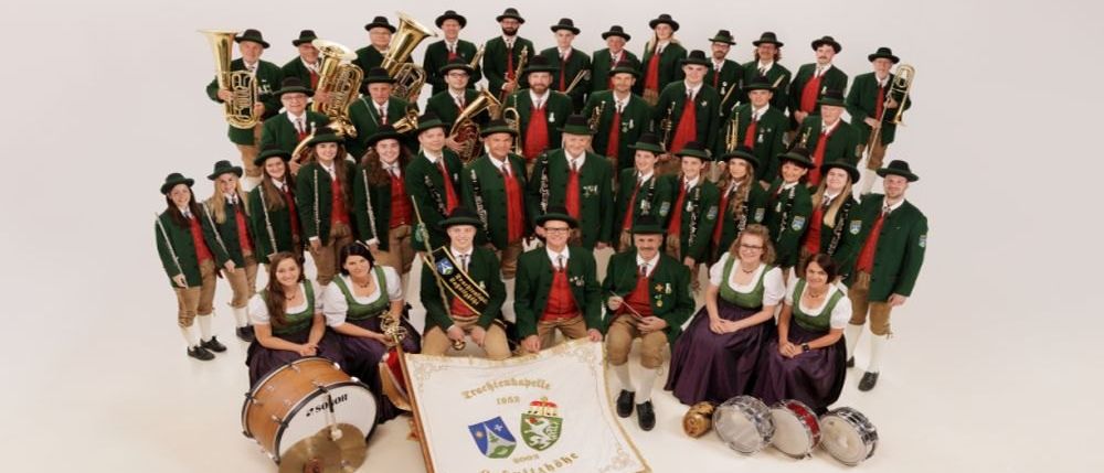 Eine große Gruppe von Musikern in grünen und roten Uniformen mit Hüten posiert für ein Foto. Einige halten Instrumente. Ein Banner mit Text und Logos wird von einer Person gehalten.