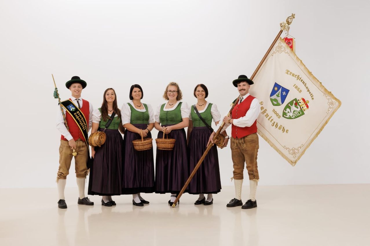Fünf Personen in traditioneller Kleidung, darunter ein Mann mit einer Flagge, posieren für ein Foto vor weißem Hintergrund.