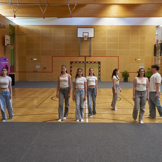 Mehrere junge Frauen in weißen Crop-Tops und Jeans stehen lächelnd auf einem Basketballfeld. Ein Basketballkorb befindet sich an der Wand hinter ihnen.