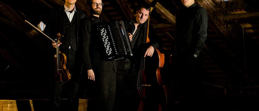 Fünf Männer in formeller Kleidung stehen in einem Dachboden und halten Instrumente: Geige, Cello und Akkordeon. Sie scheinen Musiker zu sein, die möglicherweise für ein Foto posieren.