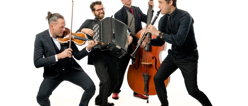 Fünf Musiker in Anzügen und Krawatten spielen Geige, Akkordeon und Klarinette, während ein Cellist mit einem Bogen steht, alle leicht geduckt in einem Studio.