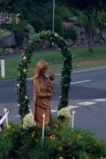Eine Statue von Maria, die ein Kind hält, ist von Grünpflanzen, Blumen und brennenden Kerzen am Straßenrand umgeben.