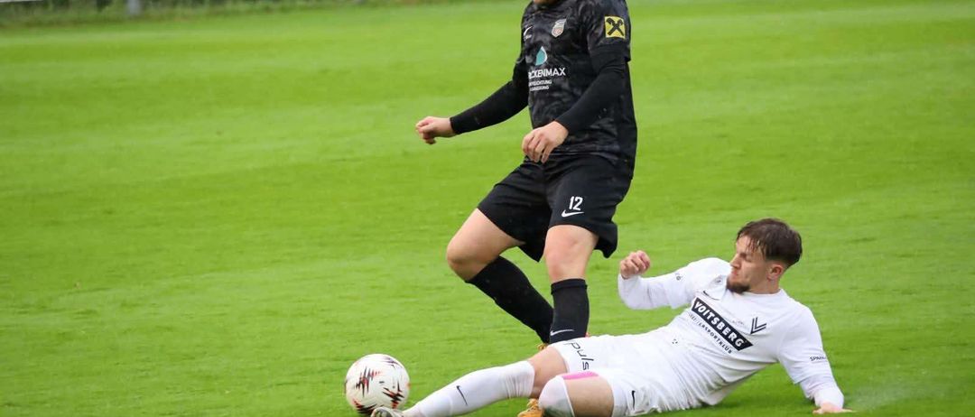 Zwei Fußballspieler sind auf einem Feld, einer tackelt den anderen. Beide tragen Trikots mit Logos. Der Spieler in Schwarz hat 'Nike' auf seinen Shorts.