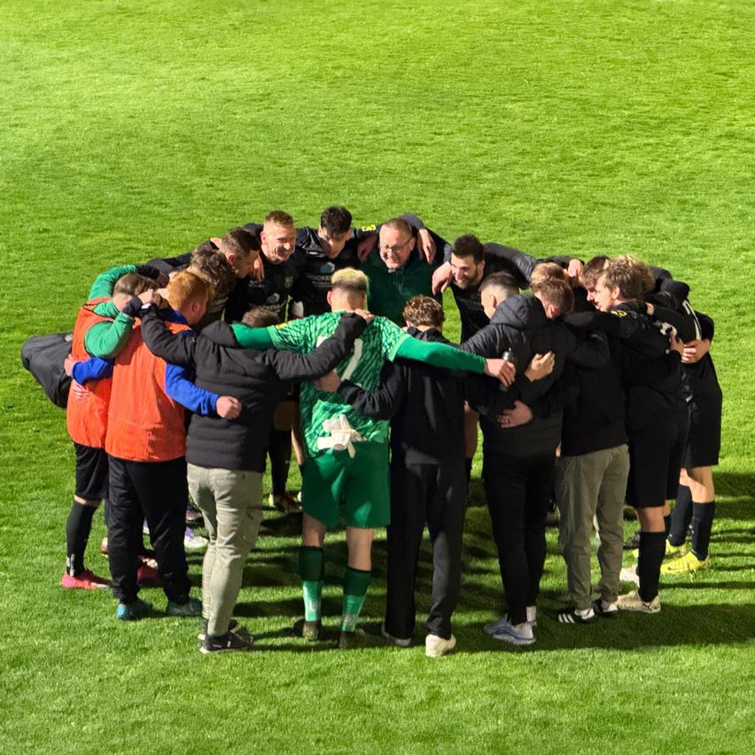 A soccer team gathers in a circle on the field, wearing green and black uniforms. They are likely preparing for a game.