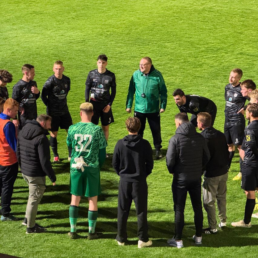 A soccer team gathers on the field, wearing black and green uniforms. A man in green stands at the center, while others listen attentively.