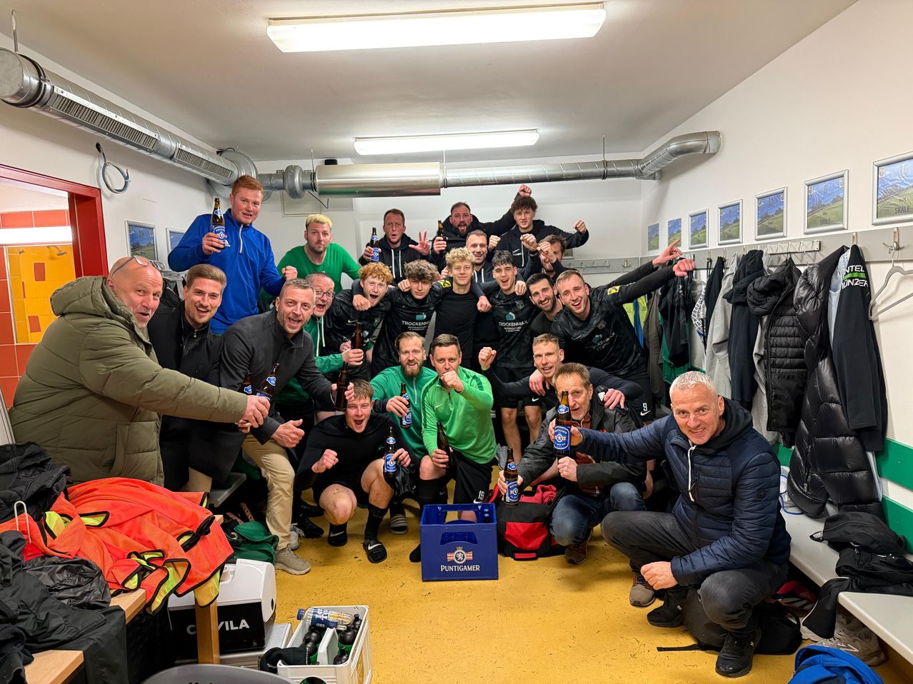 A group of soccer players celebrate in a room with beer bottles and a blue crate labeled 'PUNTIGAMER'. They are wearing jerseys and sneakers, and some are holding bottles.