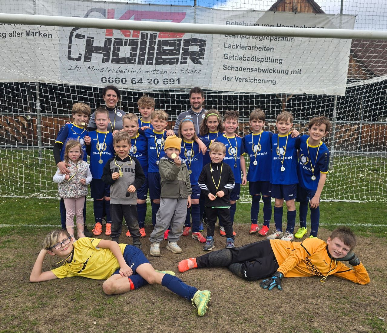 Eine Fußballmannschaft von Kindern in blauen Trikots, zusammen mit zwei Erwachsenen, posiert für ein Foto vor einem Tor mit einem Banner dahinter. Das Banner trägt den Schriftzug 'CHAUDER' und eine Telefonnummer.