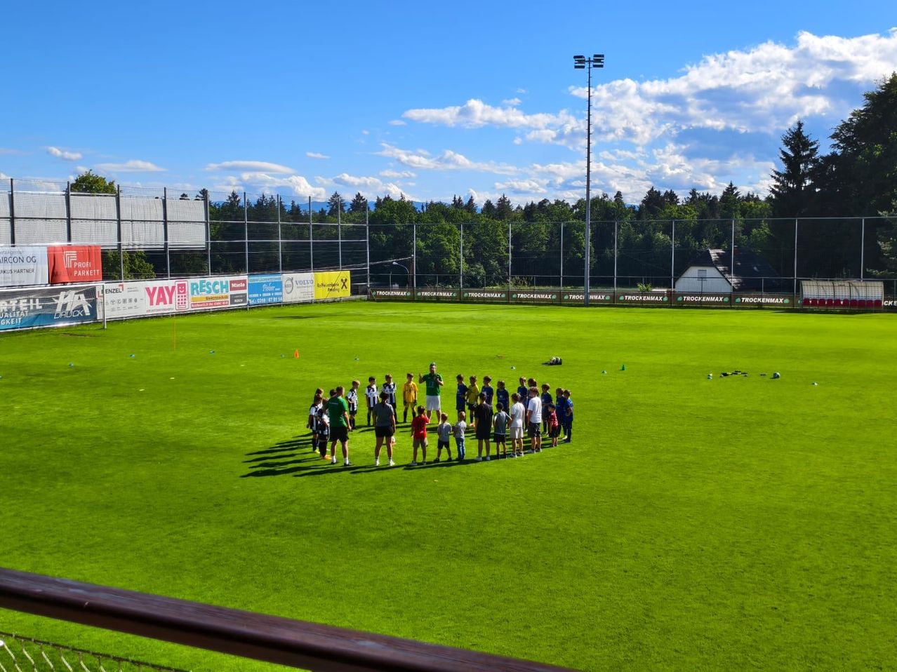 Eine Gruppe von Menschen, wahrscheinlich eine Fußballmannschaft, ist auf einem Feld versammelt. Sie sind von mehreren Werbebannern umgeben. In der Ferne steht ein großer Pfahl mit einer Lampe oben.