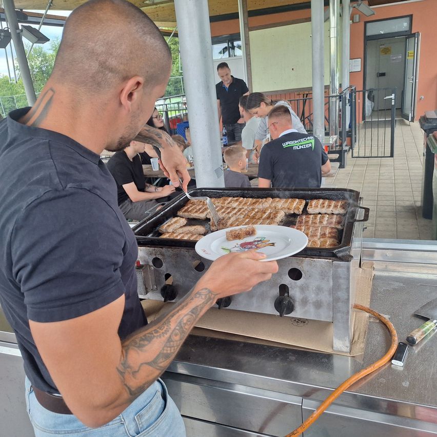 A man with tattoos is grilling food on a grill with a child and other people watching.