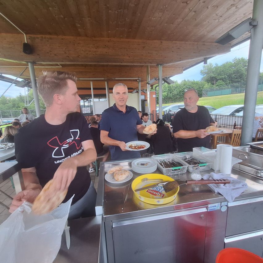 Three men in an open-air restaurant, one holding food, another holding a plate with food, and a third holding a sandwich. A table has plates, bowls, and other items.