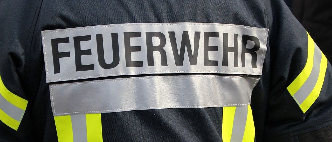 Das Feuerwehruniform zeigt ein weißes Abzeichen mit schwarzem Text 'FEUERWEHR'. Die Jacke hat reflektierende gelbe und graue Streifen.