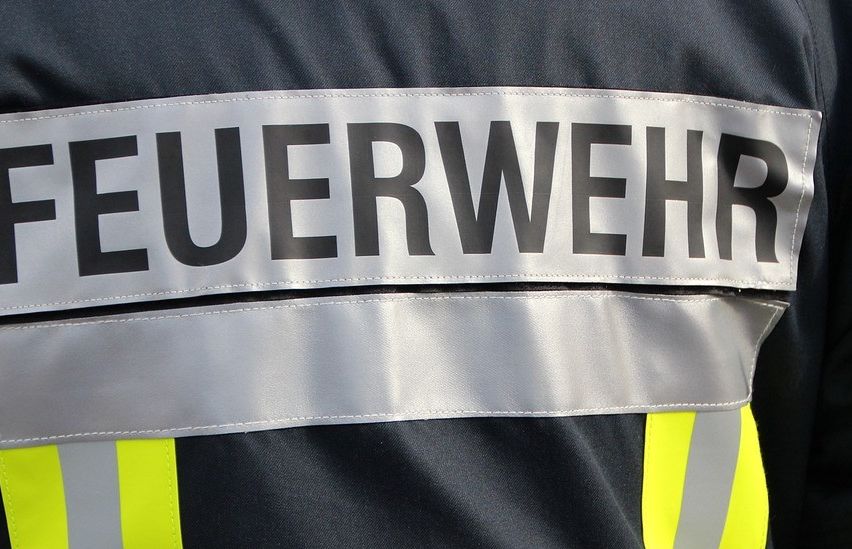 Das Feuerwehruniform zeigt ein weißes Abzeichen mit schwarzem Text 'FEUERWEHR'. Die Jacke hat reflektierende gelbe und graue Streifen.