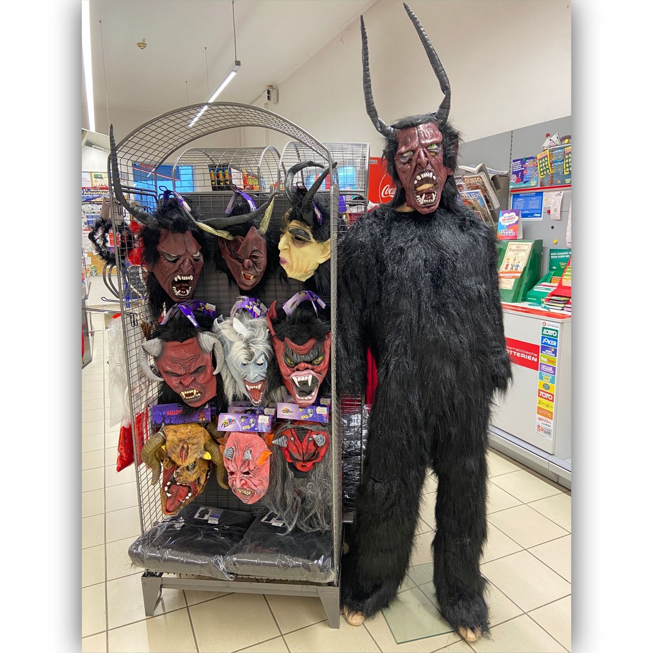 Eine als Teufel verkleidete Person steht neben einer Ausstellung verschiedener Teufelsmasken in einem Geschäft.