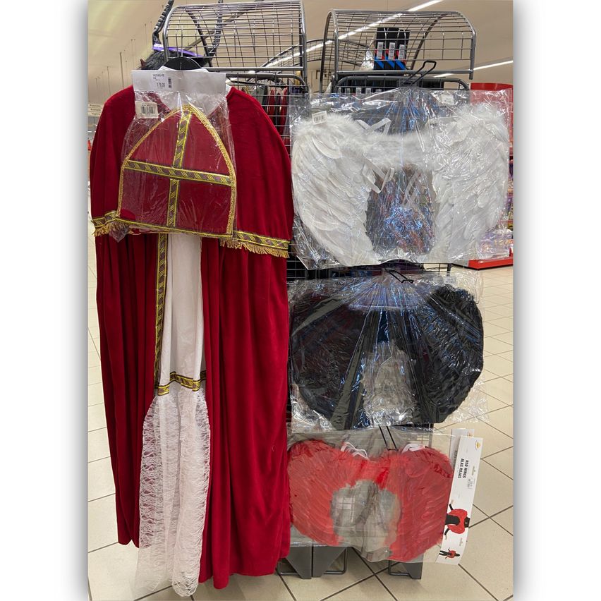 Ein roter Umhang mit weißem Spitzeninnenfutter und einem roten Hut mit goldenem Besatz ist auf einem Ständer ausgestellt. In der Nähe sind auch Engelsflügel und ein rotes Kostüm zu sehen.