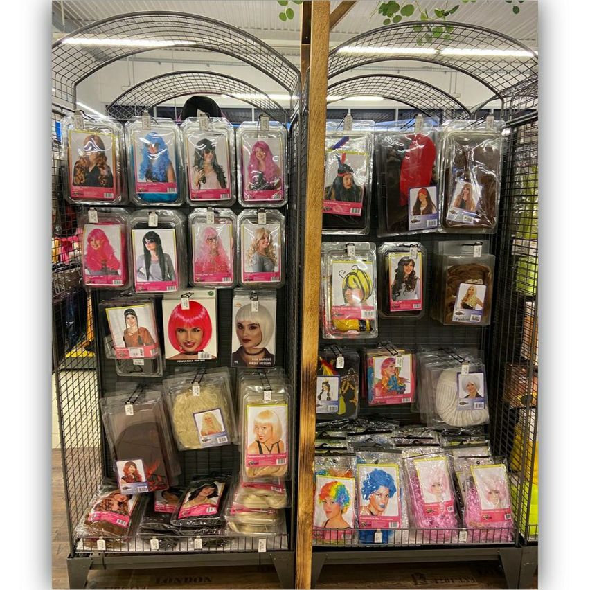 A store shelf displays a variety of wigs and hairpieces, each in plastic packaging with images of women with different hairstyles. The products range from black and blonde to vibrant pink and multicolored wigs.