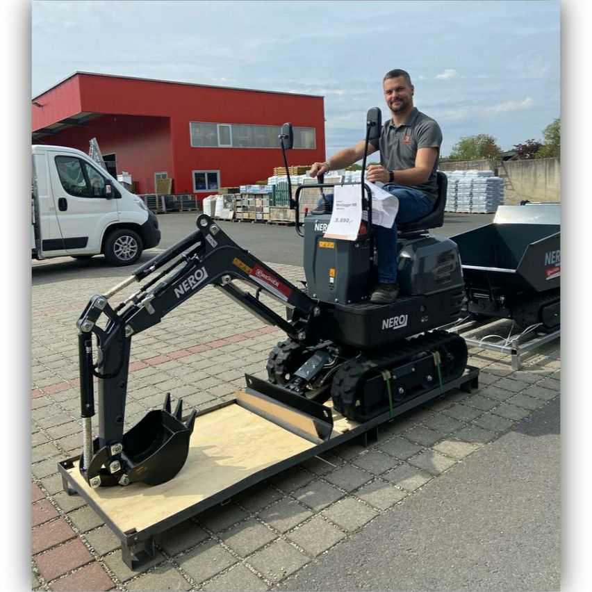 Ein Mann sitzt auf einem schwarzen Minibagger mit dem Wort NerO darauf und hält ein Stück Papier in der Hand. Dahinter steht ein weißer Lieferwagen und ein rotes Gebäude.