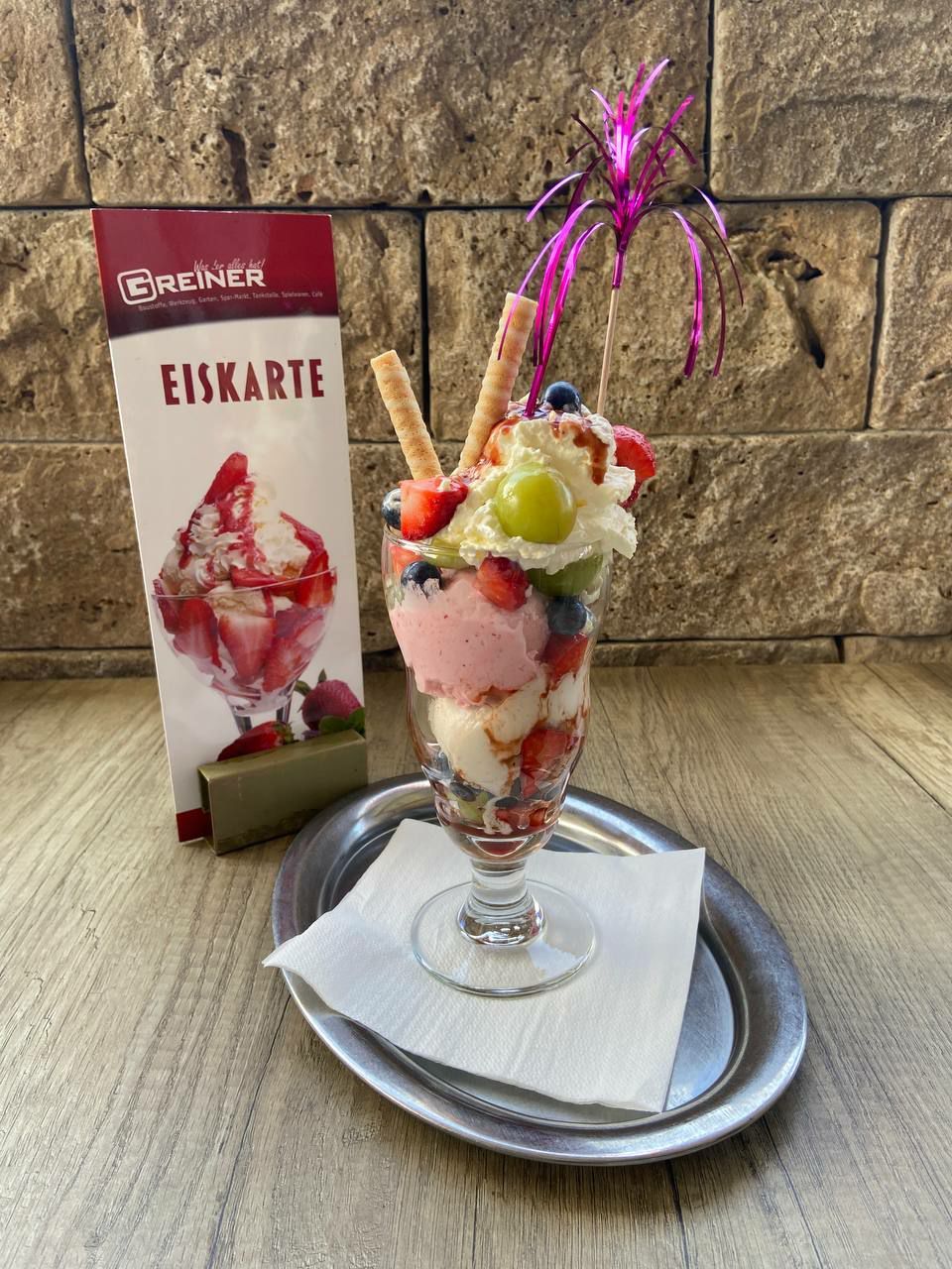 Ein Glas mit einem mehrschichtigen Dessert aus Eiscreme, Früchten und Schlagsahne, gekrönt von einem lila Papierkegel, auf einem silbernen Teller mit einer Serviette, neben einem Menükarten.