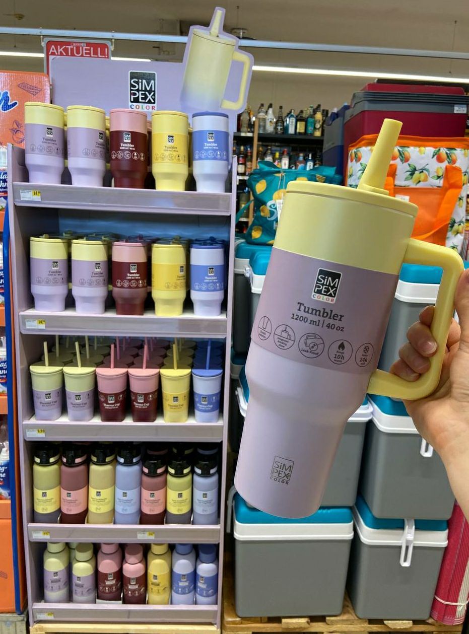 Eine Person hält einen gelben und lilafarbenen Becher mit einem Strohhalm. Dahinter zeigt ein Regal mehrere Becher in verschiedenen Farben und Größen.