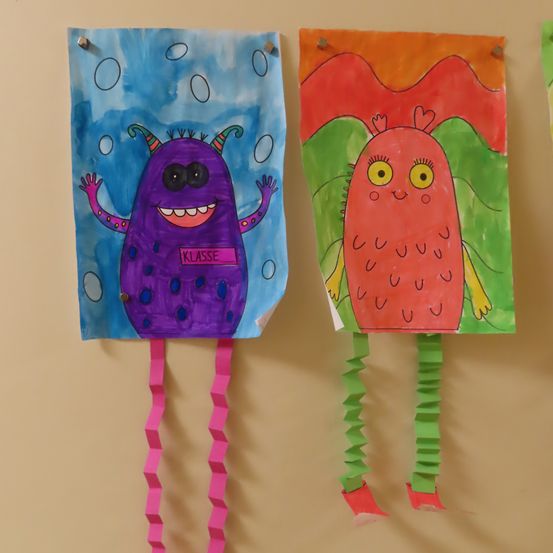 Zwei farbenfrohe Papierausschnitte von Monstern hängen an einer hellgelben Wand. Links ist ein lilafarbener mit einem Schild, das 'KLASSE' steht. Rechts ist ein roter mit grünen Akzenten und gelben Augen.