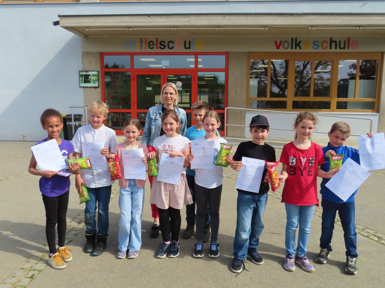 Eine Gruppe von Schülern einer Mittelschule mit einer Lehrerin steht vor einem Gebäude, hält Urkunden und Snacks in der Hand.