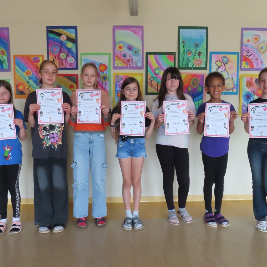 Sieben Mädchen mit verschieden farbiger Kleidung und Schuhen stehen in einem Raum mit Holzboden. Sie halten ein Zertifikat in den Händen und lächeln.