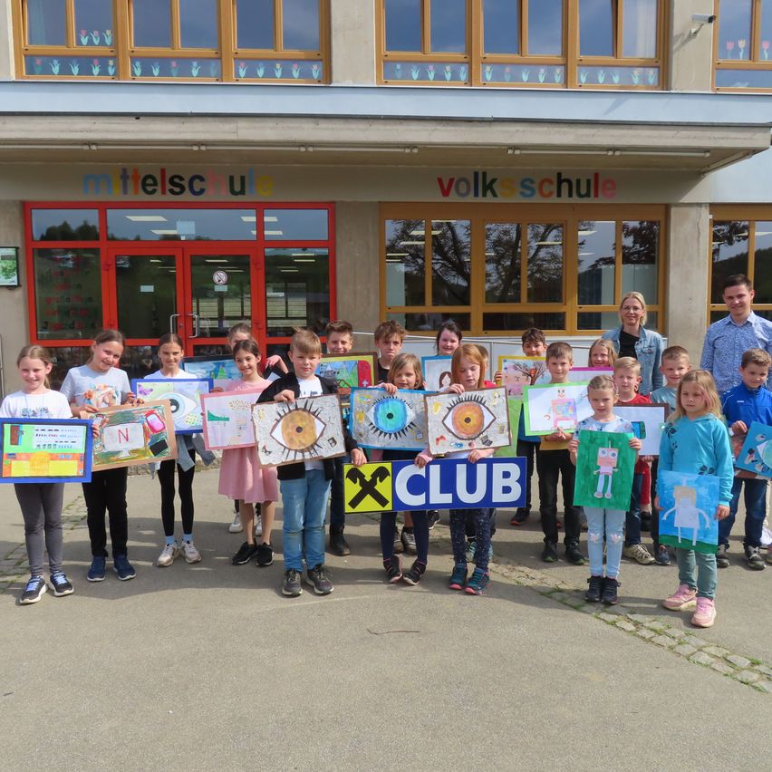Eine Gruppe von Kindern mit farbenfrohen Kunstprojekten steht vor einem Schulgebäude mit dem Schild 'Mittelschule Volksschule'.