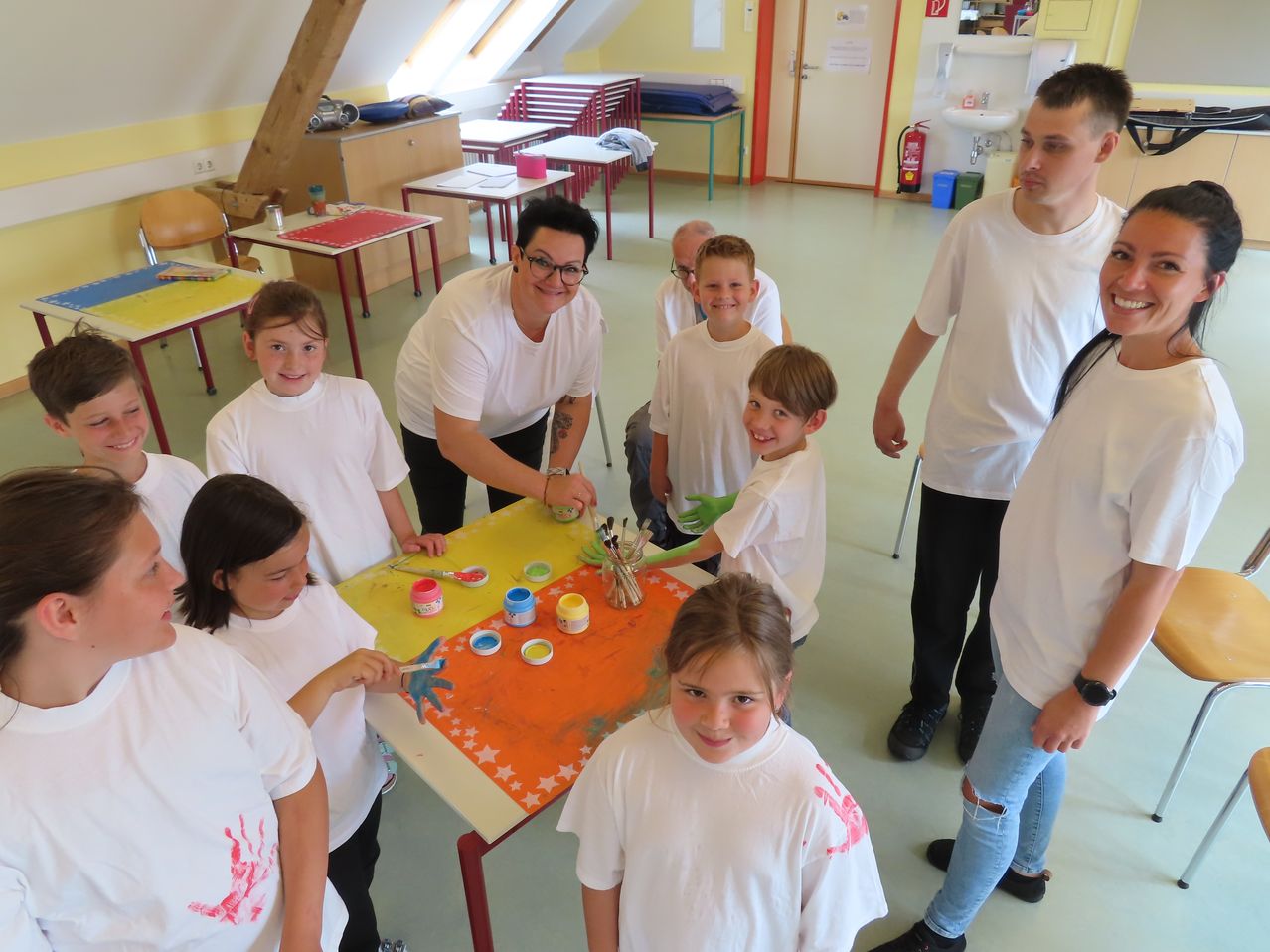 Eine Gruppe von Kindern in weißen T-Shirts malt einen Tisch, während eine Frau mit Brille hilft. Mehrere Erwachsene stehen um sie herum. Der Raum hat gelbe Wände und eine Holzdecke.