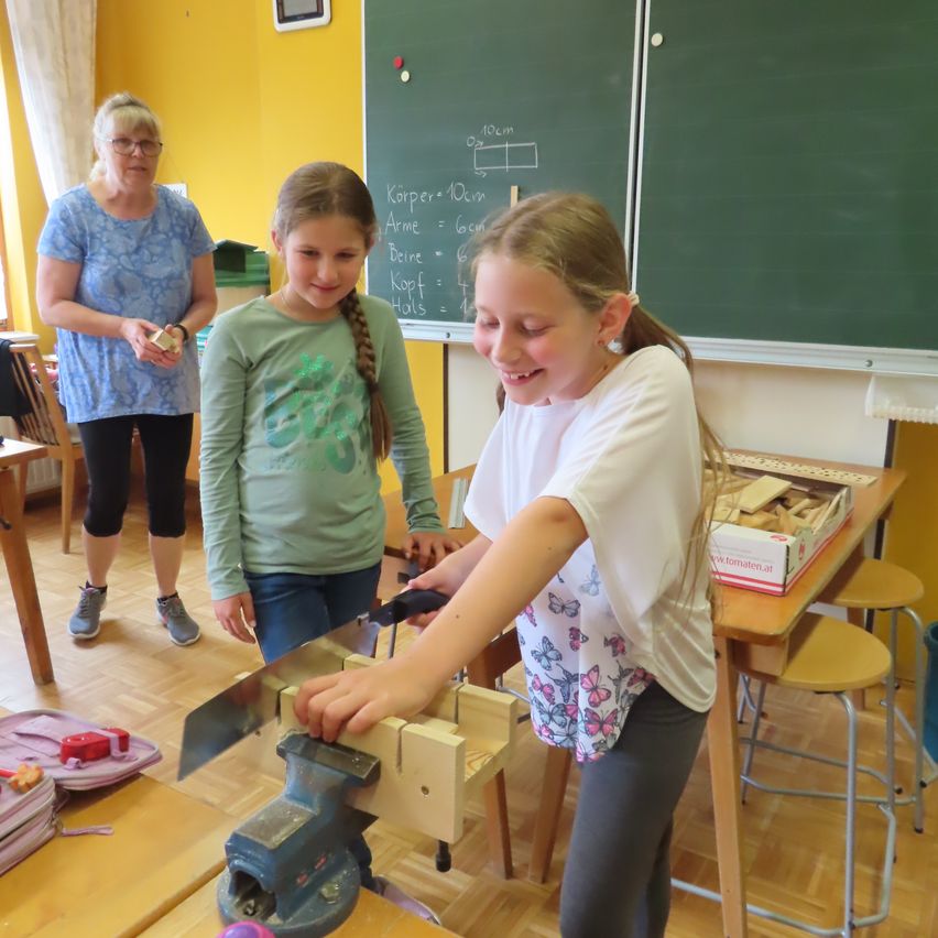 Ein Mädchen benutzt eine Säge in einem Klassenzimmer, während ein anderes Mädchen und eine Frau zusehen. Ein Tafel mit mathematischen Gleichungen ist hinter ihnen.
