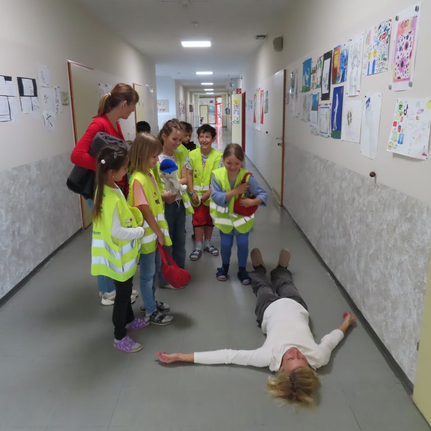 In einem Flur liegt eine Frau auf dem Boden, während mehrere Kinder in Sicherheitswesten um sie herumstehen, und eine andere Frau zusieht. An den Wänden hängen Zeichnungen.