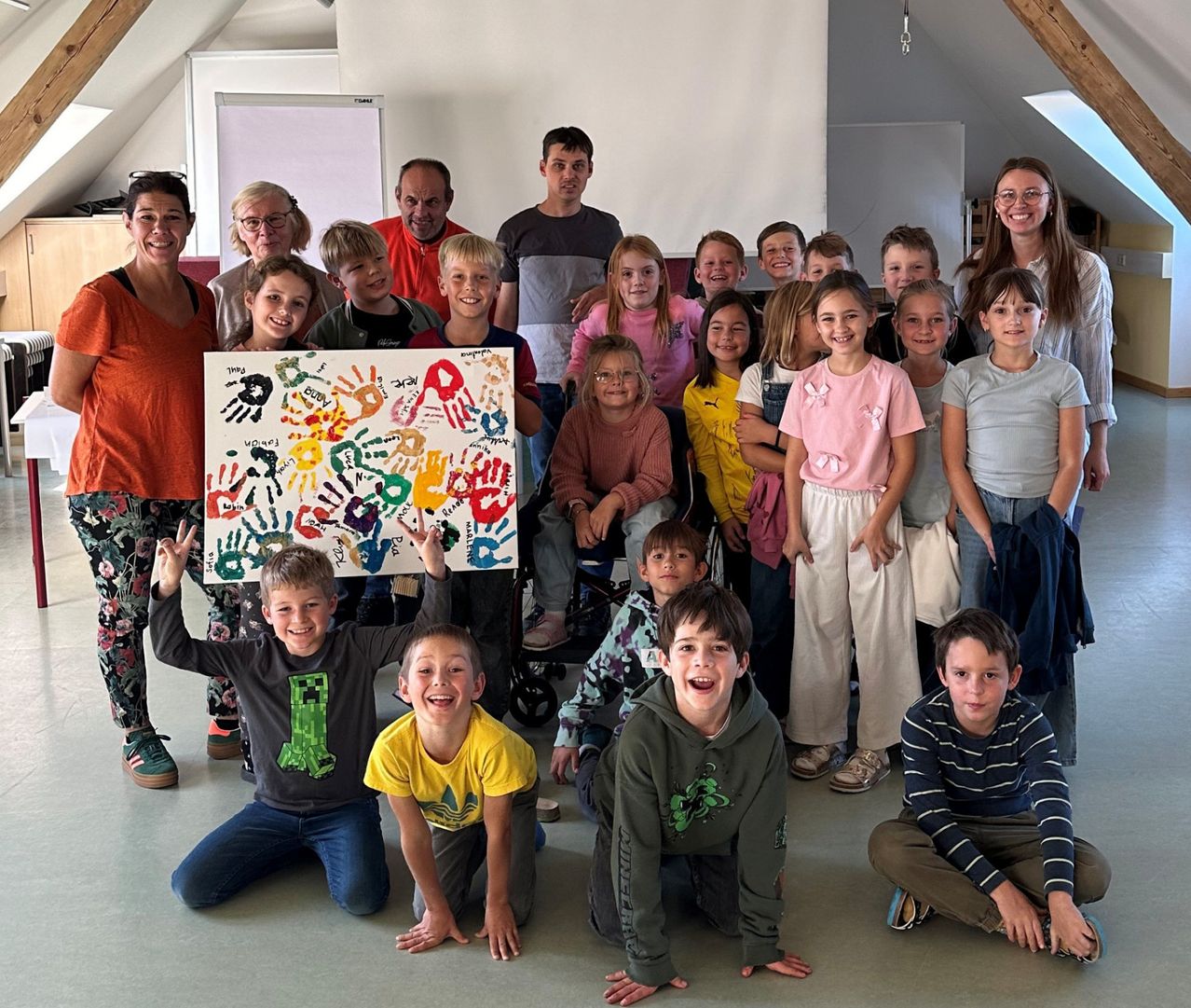 Eine Gruppe von Kindern und Erwachsenen posiert für ein Foto mit einer farbenfrohen Handabdruck-Leinwand, lächelnd in einem Raum mit weißen Wänden und einer Holzdecke.