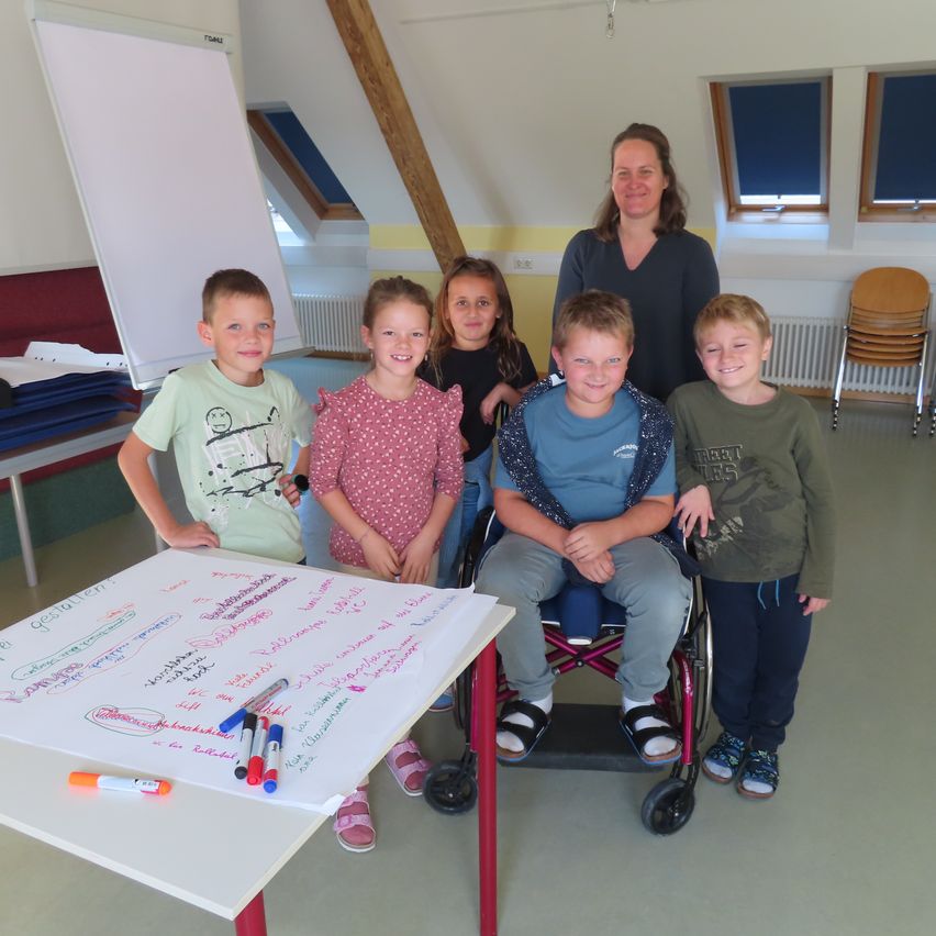 Eine Gruppe von Kindern und einer Frau steht um einen Tisch mit einer Tafel in einem Klassenzimmer. Die Kinder lächeln und posieren für ein Foto. Die Tafel hat Zeichnungen und Schriften. Die Frau steht neben einem Kind im Rollstuhl. Dahinter sind Fenster, Stühle und ein Heizkörper.