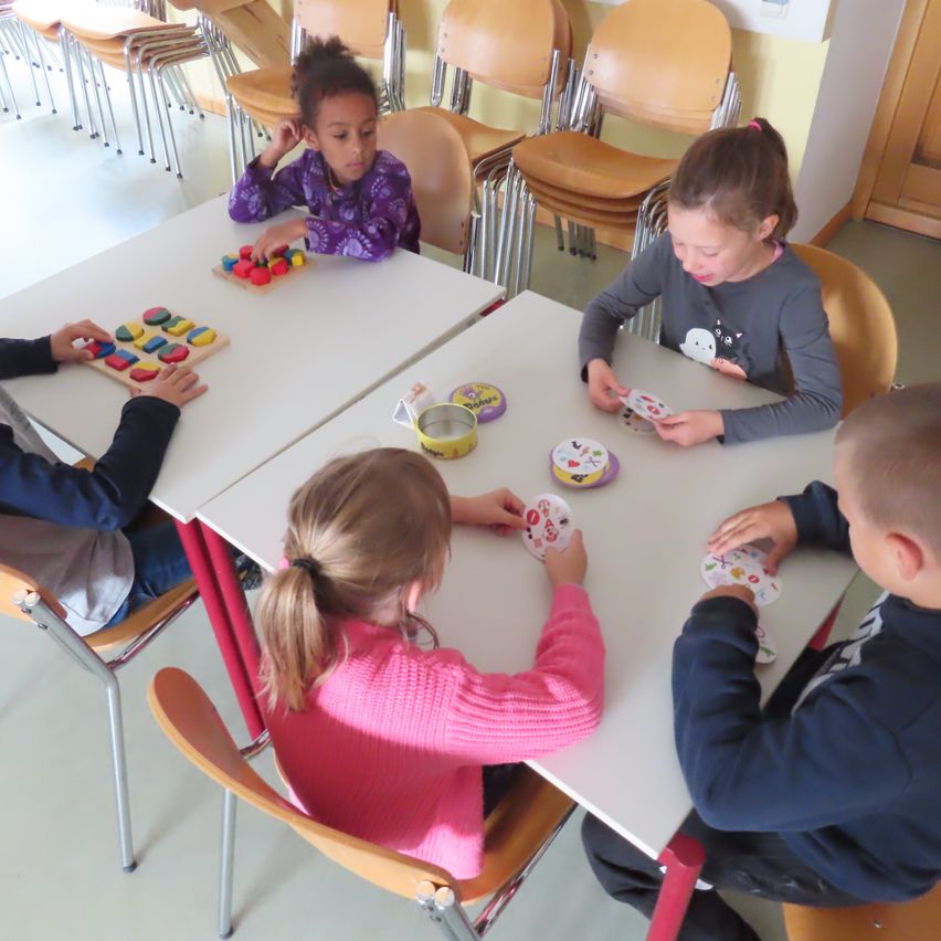 Fünf Kinder sitzen um einen Tisch herum und spielen mit Spielzeug. Auf dem Tisch befinden sich Karten und ein Holzpuzzle. Die Kinder scheinen in das Spiel vertieft zu sein.