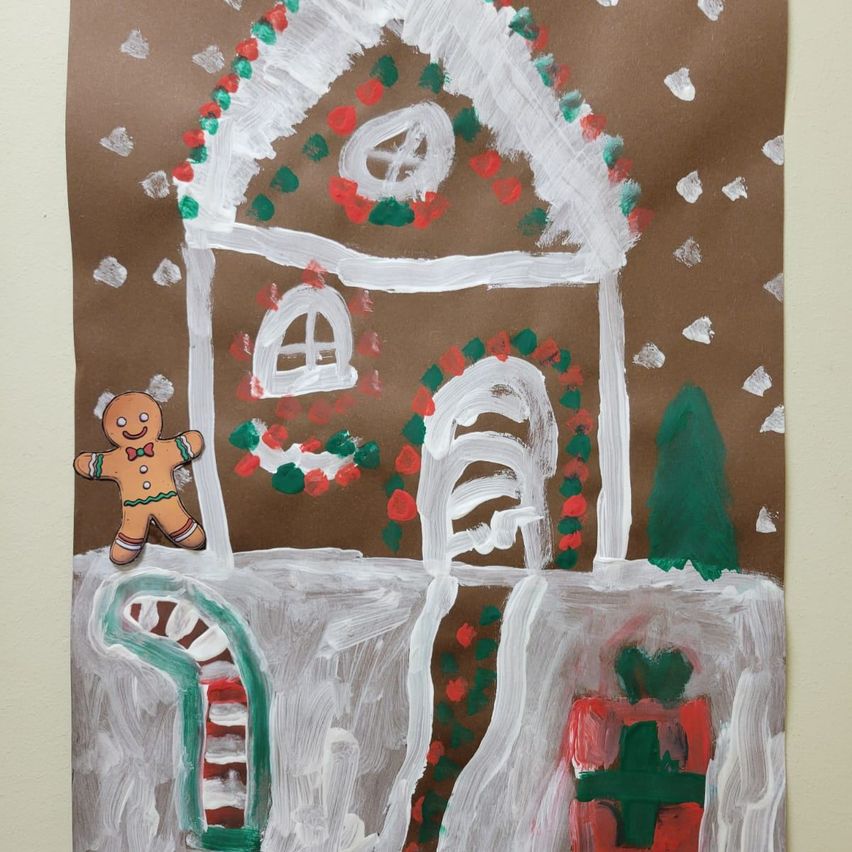 A painted gingerbread house with a snow-covered roof, decorated with red and green candies. A gingerbread man stands in front of it, with a candy cane nearby.