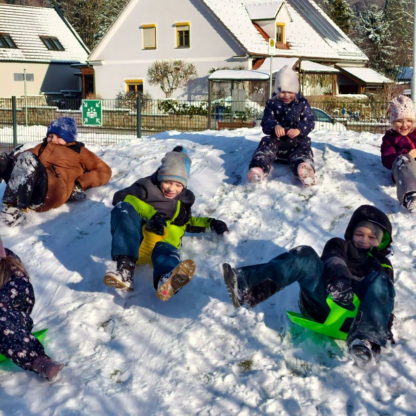 Fünf Kinder spielen im Schnee. Sie sitzen auf Schlitten, und einige stehen. Sie alle lächeln und scheinen den Schnee zu genießen.