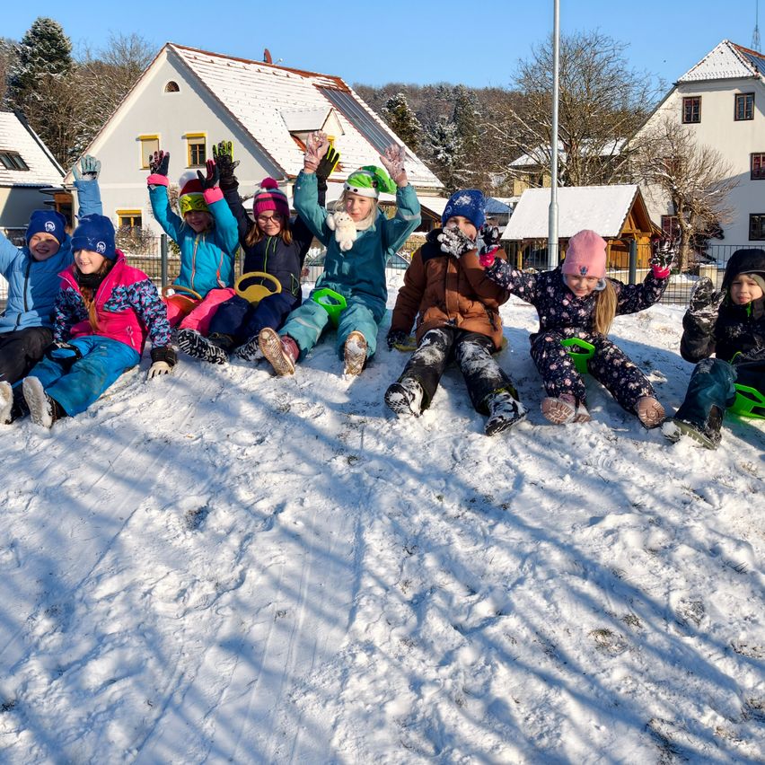 Eine Gruppe von Kindern in Winterkleidung sitzt im Schnee, einige mit Handschuhen hoch. Sie lächeln und posieren für ein Foto. Hinter ihnen stehen Häuser mit schneebedeckten Dächern. Die Häuser sind von Bäumen umgeben.