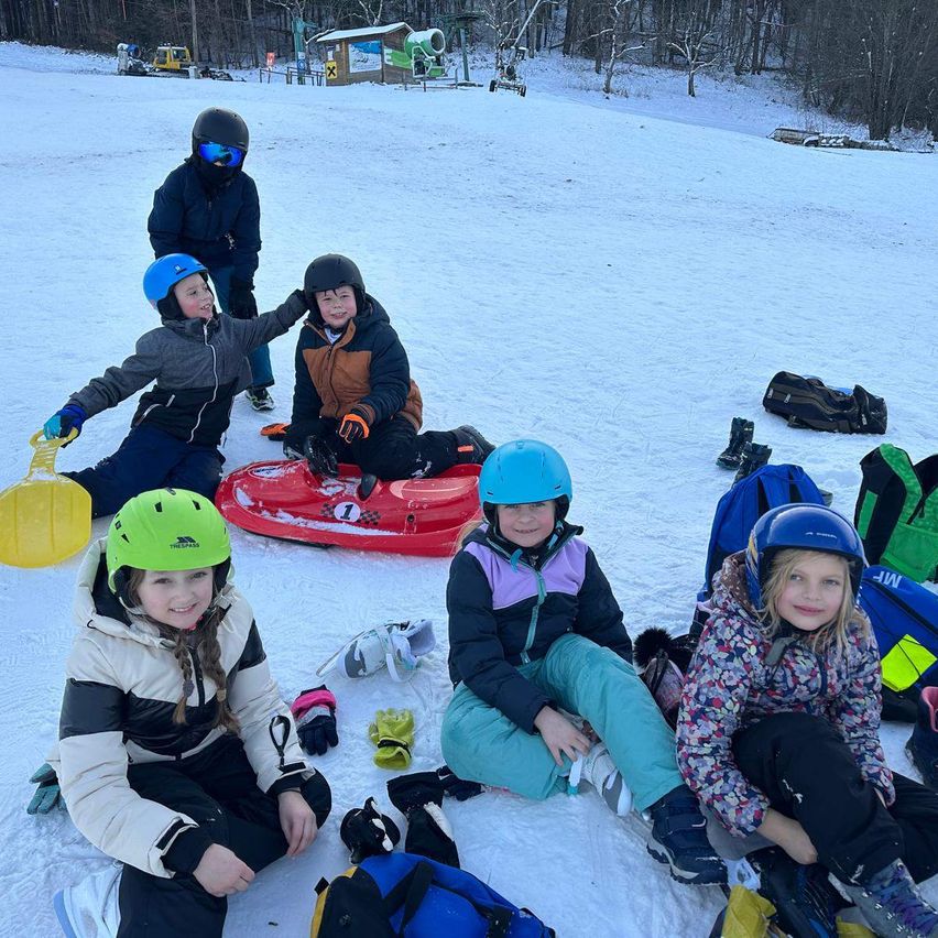 Eine Gruppe von Kindern in Winterkleidung sitzt im Schnee. Sie tragen Helme und Handschuhe. Einer von ihnen hält einen Schlitten, und es gibt Taschen und Schlitten auf dem Boden. Bäume und ein Gebäude sind im Hintergrund.