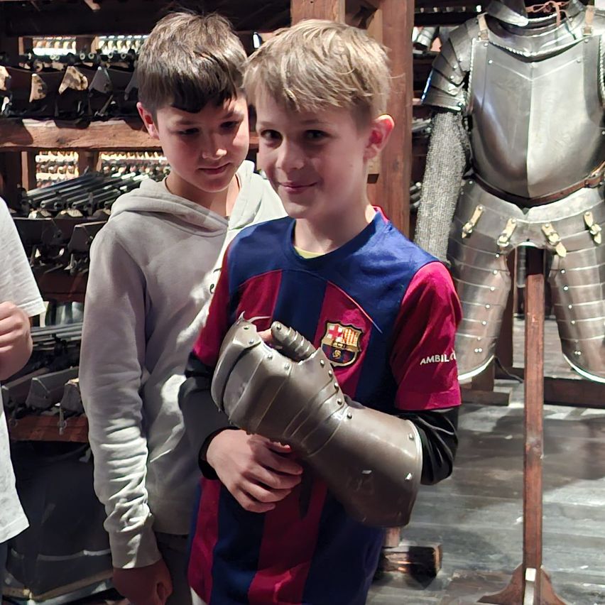 Two boys stand next to a medieval knight's armor. One boy wears a Barcelona soccer jersey and holds a knight's gauntlet.
