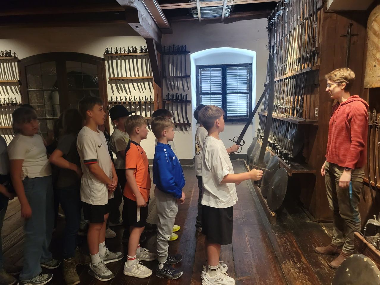 A group of children are standing in a room with wooden floors and walls, observing a sword and shield held by a boy in front.