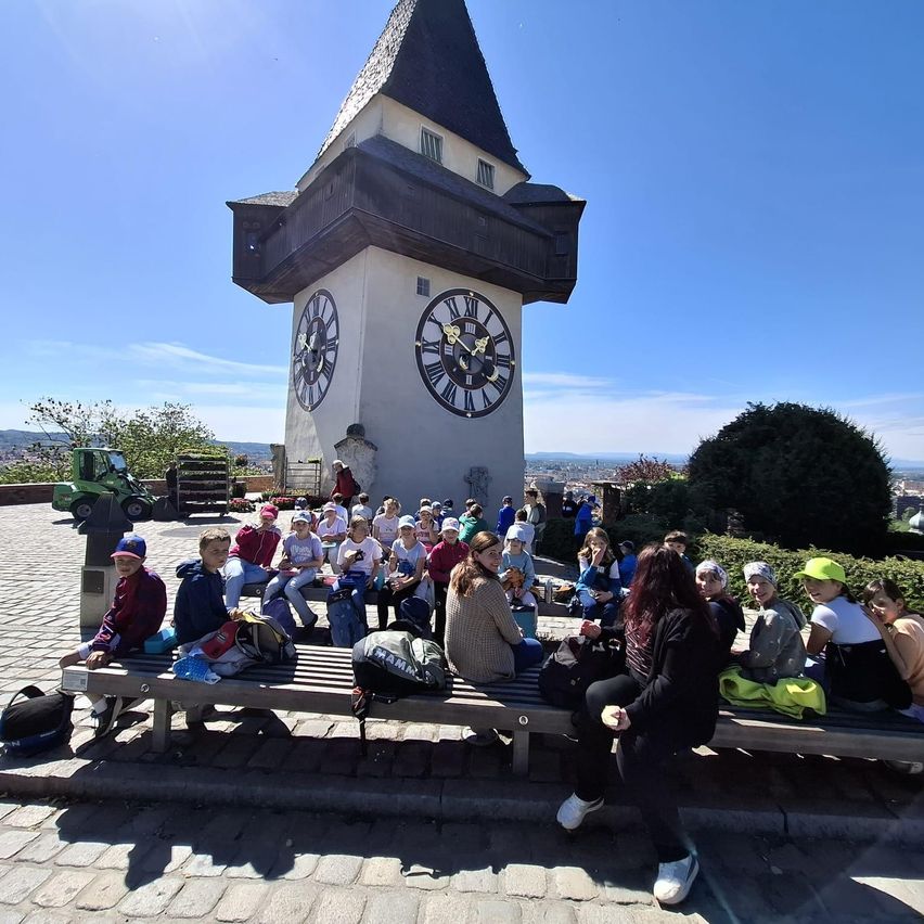 A group of people are seated on benches in front of a tower clock with two faces, under a clear sky.