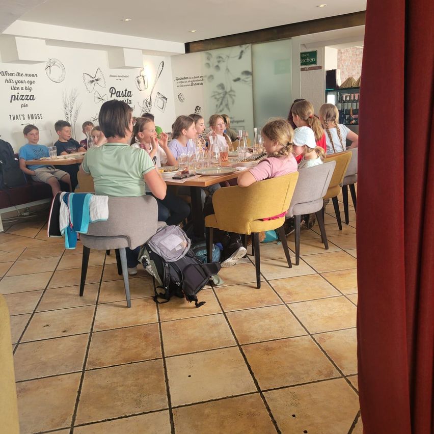 A group of children are sitting at tables inside a restaurant, eating and drinking. The restaurant has tiled floors and walls with drawings. Backpacks and plates are on the tables.