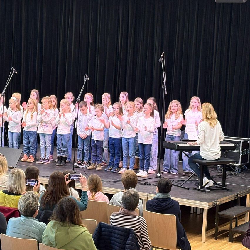 Ein Chor junger Kinder tritt mit einem Pianisten auf der Bühne auf, während ein Publikum zuschaut. Die Kinder stehen in ordentlichen Reihen, und einige Erwachsene machen Fotos.