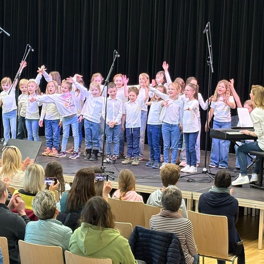 Eine Gruppe von Kindern in weißen Hemden und Jeans steht auf der Bühne und singt. Sie heben die Hände und lächeln. Hinter ihnen steht ein Klavier, das eine Frau spielt. Davor sitzen Erwachsene auf Stühlen und beobachten die Aufführung. Einige halten ihre Handys, um sie aufzuzeichnen.