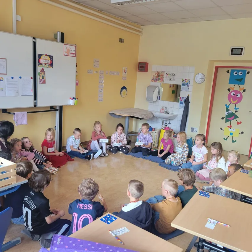 Ein Klassenzimmer mit Kindern, die im Kreis sitzen, einer Lehrerin, die in der Nähe steht, und verschiedenen pädagogischen Postern und Dekorationen an den Wänden.